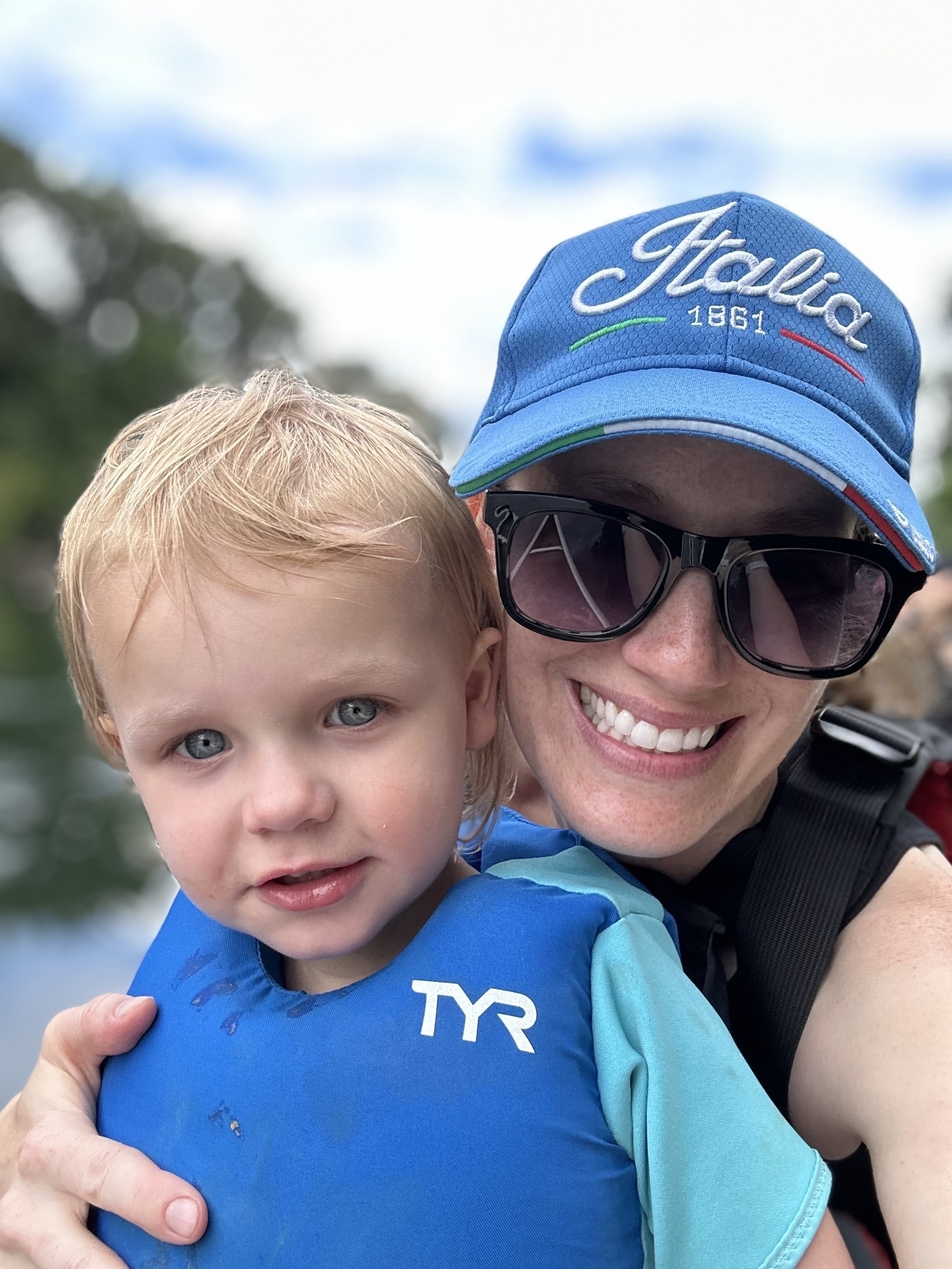 A smiling adult wearing sunglasses and a blue cap is holding a young child who is wearing a blue TYR shirt.