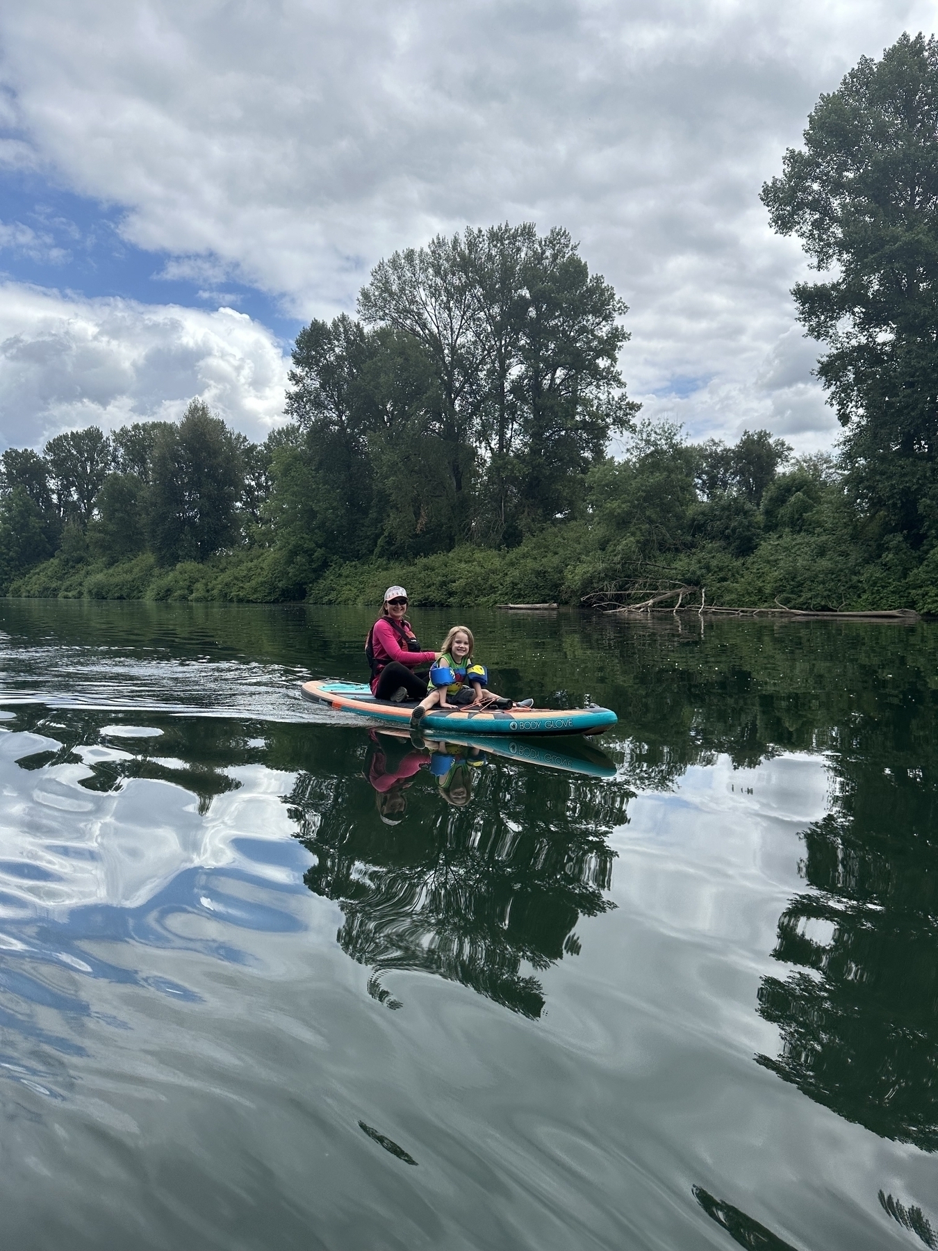 Two people are sitting on a paddleboard floating on a calm, reflective body of water surrounded by lush greenery and trees under a cloudy sky.